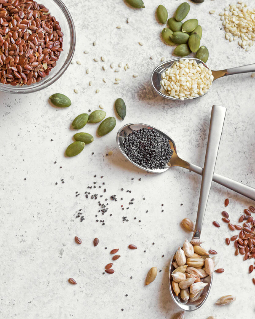 Assorted seeds and grains on a light surface with spoons.
