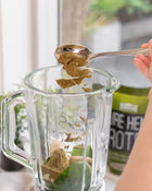 Brown hemp protein powder being added with a spoon to a glass blitzer with spinach leaves.