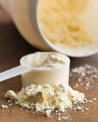 White scoop with Whey protein powder on a wooden surface, with a container of powder in the background.