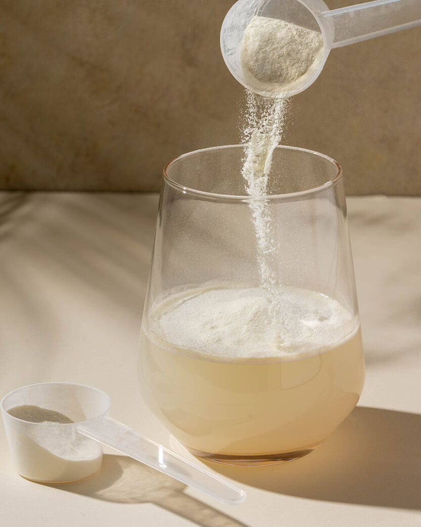 Powder being poured from a scoop into a glass of liquid on a neutral background