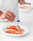 Person seasoning salmon fillets with a sauce mixture on a white plate.