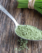 Spoonful of green powder on a wooden surface with fresh herbs in the background