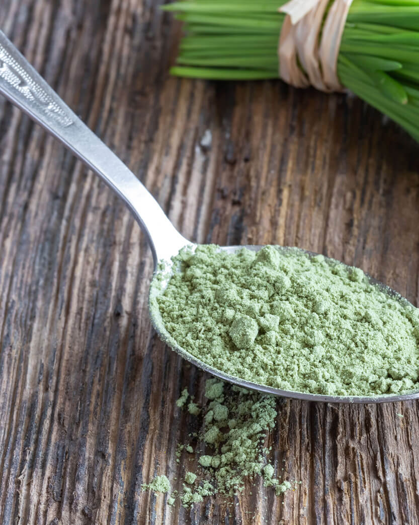 Spoonful of green powder on a wooden surface with fresh herbs in the background