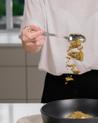 Person holding a spoon with curry powder over a black bowl in a kitchen setting