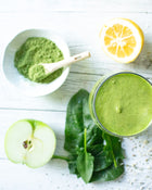 Green smoothie with ingredients including a bowl of green powder, lemon, apple, and spinach on a white surface.