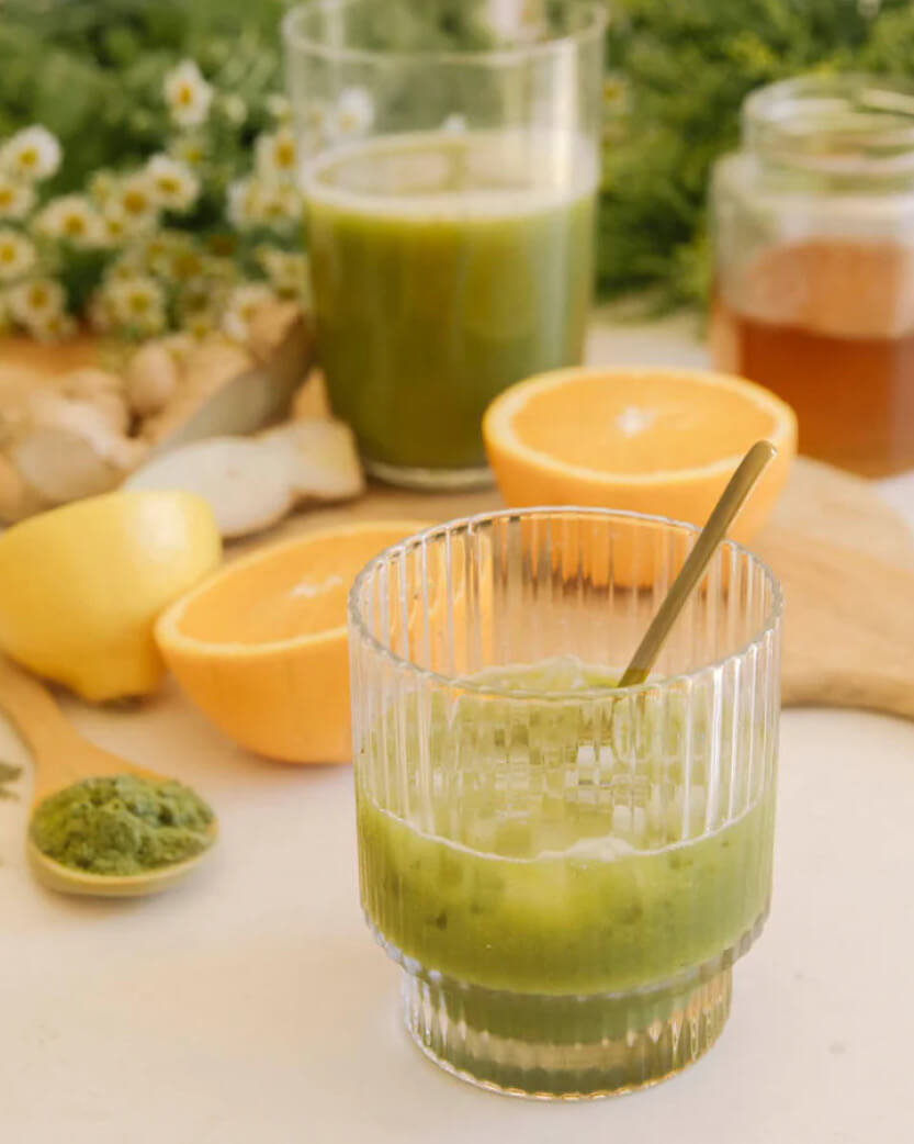 Green smoothie in a glass with a spoon, surrounded by oranges, lemons, and herbs on a light surface.