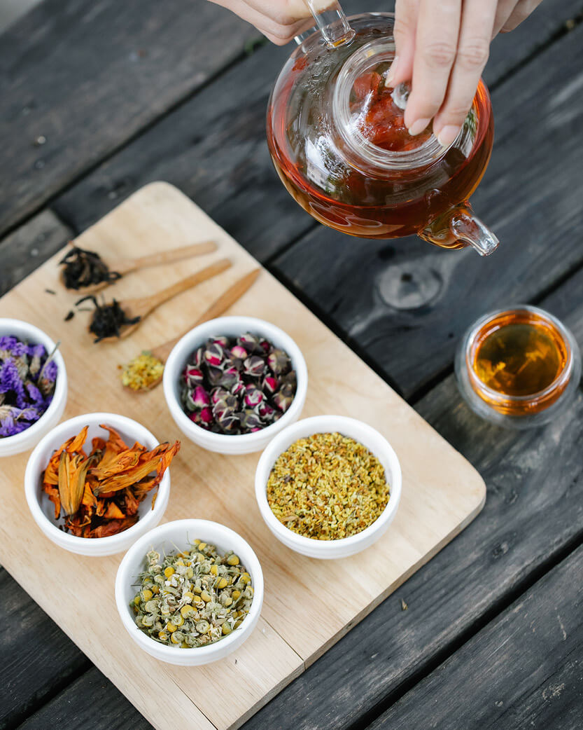bowls of dried herbs with a glass pot of tea being poured into a cup on a dark wooden table. 