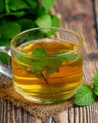Clear glass mug filled with green tea and mint leaves on a wooden surface.