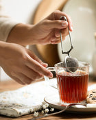 Person using a tea infuser to steep tea in a glass cup on a wooden table.