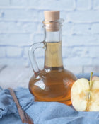 Glass bottle of apple cider vinegar with a cork lid on a blue cloth with apples.