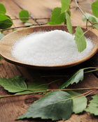 Wooden bowl filled with white granulated substance surrounded by green leaves on a wooden surface