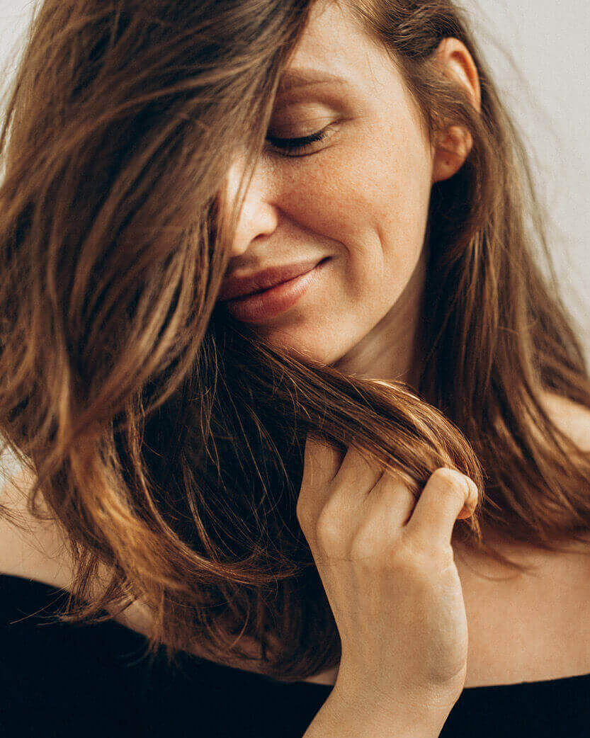 Woman with long brown hair touching her face against a neutral background.
