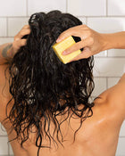 Person washing hair with a yellow sponge in a bathroom setting.