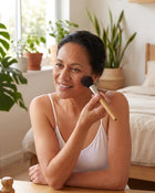Woman with dark hair applying makeup with a brush in a bedroom setting.