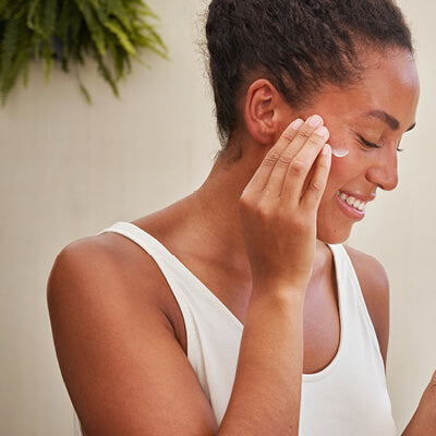 Woman applying cream to her face with a neutral background.
