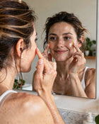Woman applying cream to her face in front of a mirror.