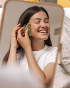 Woman using a green facial roller in front of a mirror.
