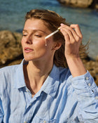 Woman applying serum to her face with a dropper by the sea.