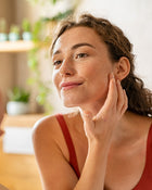 Woman in a red tank top touching her face in a softly blurred indoor setting.