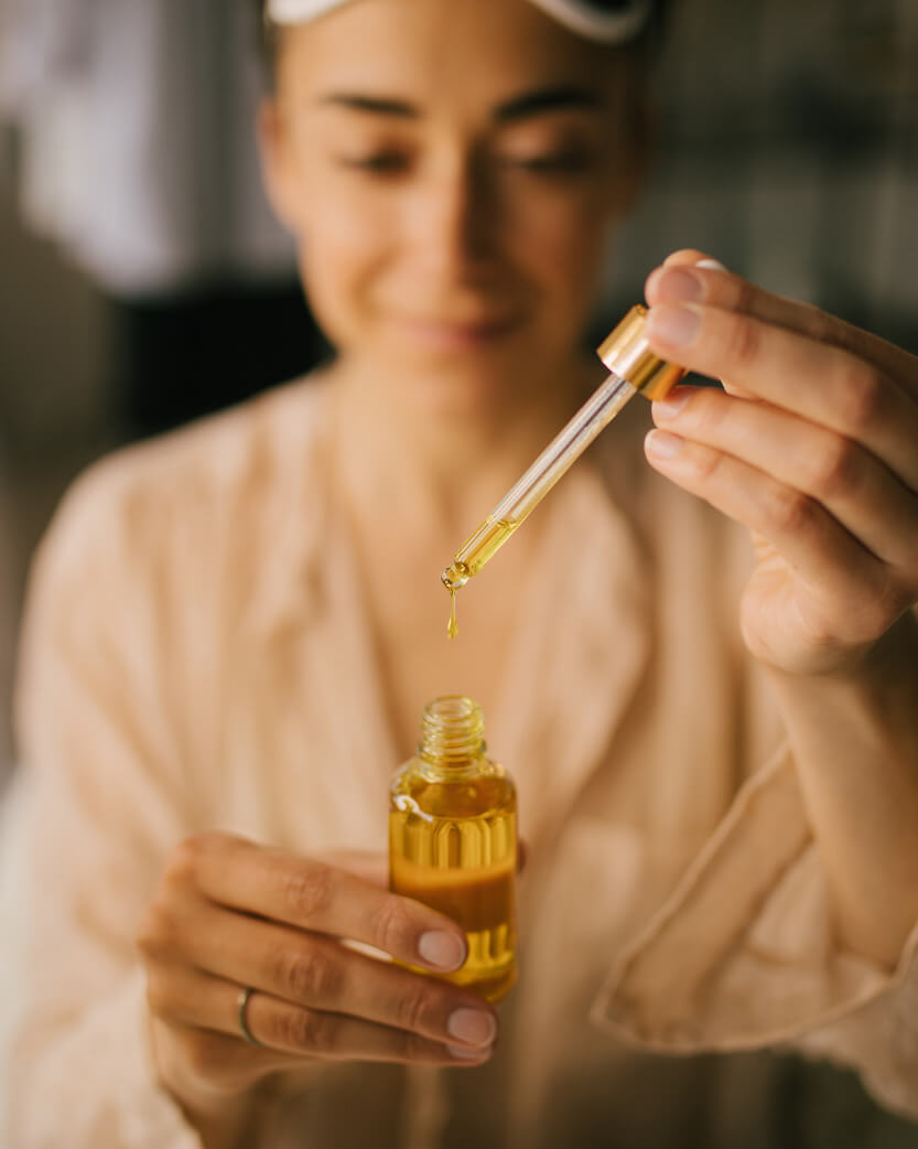 Person holding a dropper over a bottle of yellow liquid, possibly oil or serum.
