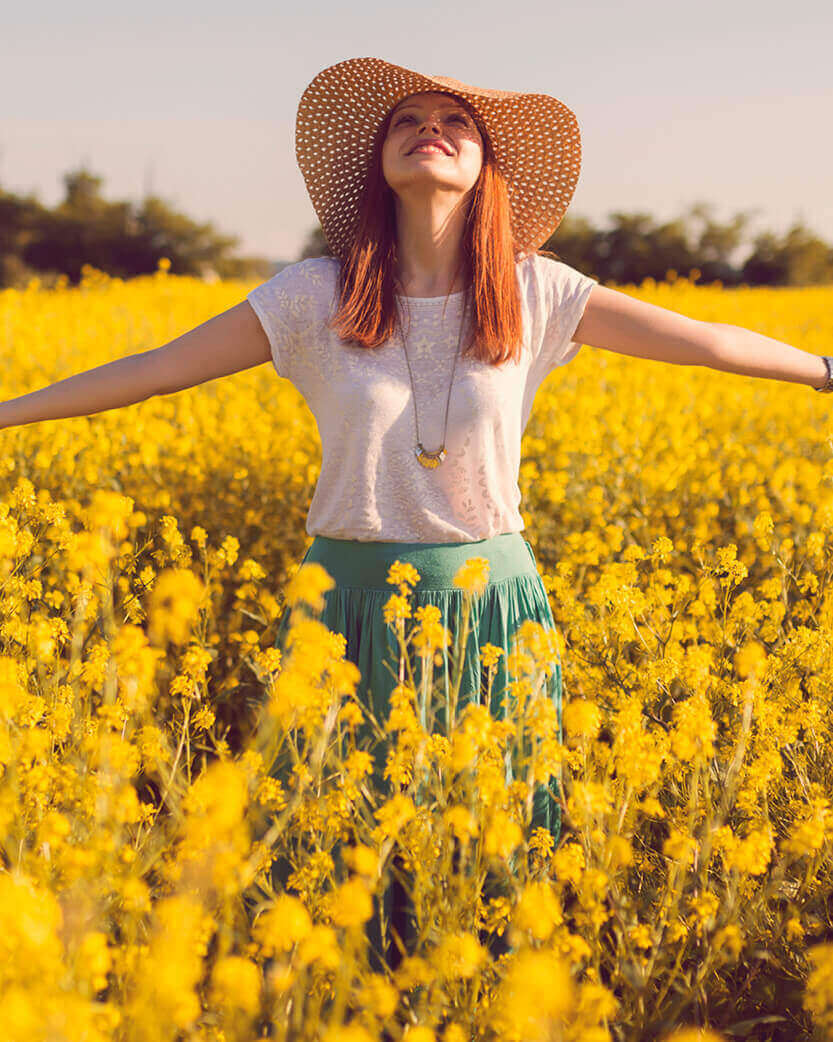 Woman in a straw hat standing in a field of yellow flowers with arms outstretched.