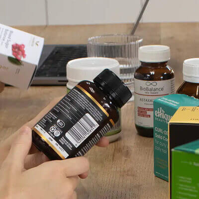 Hand holding a bottle of supplements with various other supplement bottles on a wooden surface.