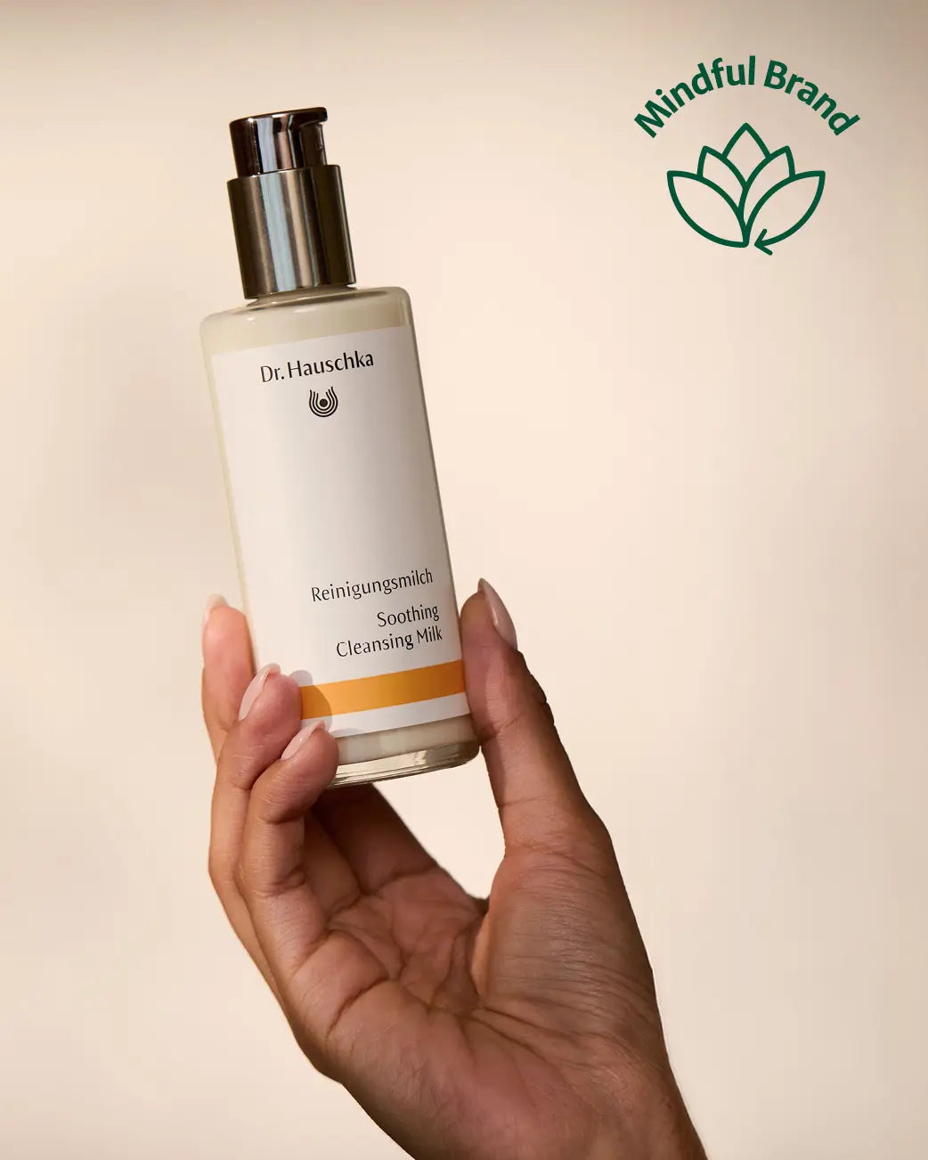 Hand holding Dr Hauschka Soothing Cleansing Milk pump bottle up against a pale cream backdrop. Green mindful brand logo in the corner.
