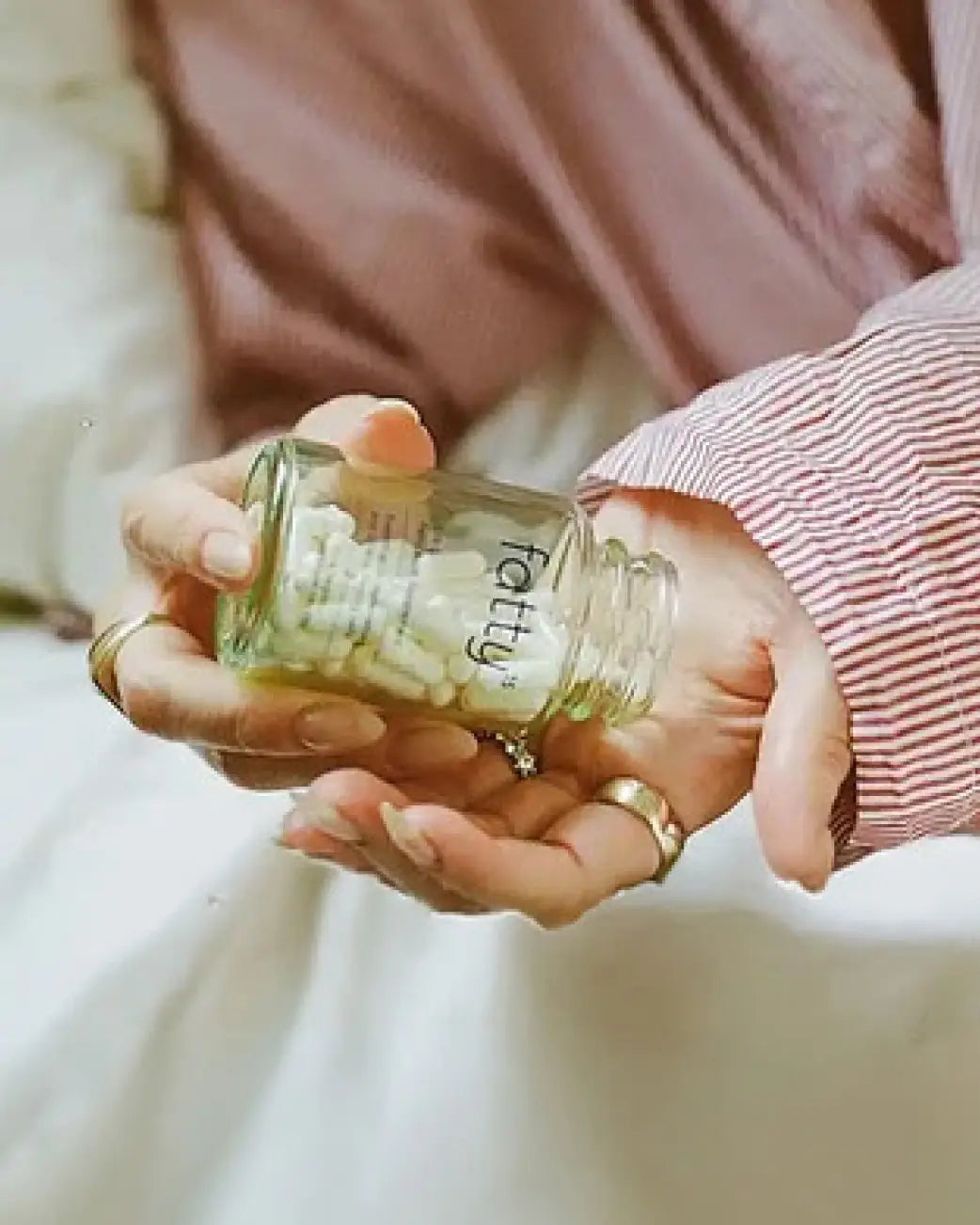A person holding a small clear glass bottle filled with white capsules, with a pink and white striped shirt visible in the background.