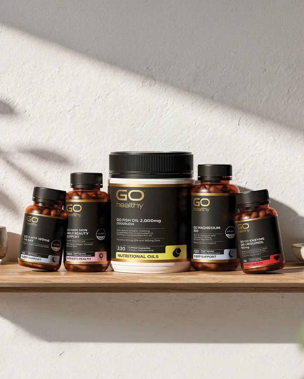 Five GO healthy dietary supplement bottles arranged on a wooden shelf against a textured white wall with soft shadow patterns. The collection includes various fish oil and essential oil products in amber bottles with black caps and black labels featuring gold and white text.