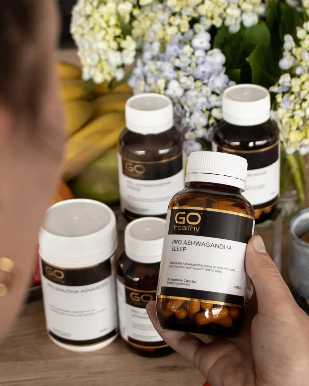 A hand holds a bottle of GO Healthy Pro Ashwagandha Sleep capsules in the foreground, with several other GO Healthy supplement bottles arranged on a wooden surface in the background, with flowers and a person partially visible.
