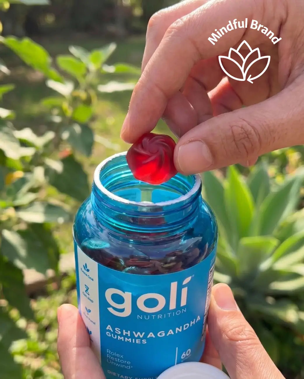 A hand holds an open blue bottle of Goli Nutrition Ashwagandha Gummies outdoors, with a red gummy being lifted from the bottle. The background shows blurred green foliage, and a 'Mindful Brand' logo watermark is visible in the top right corner.