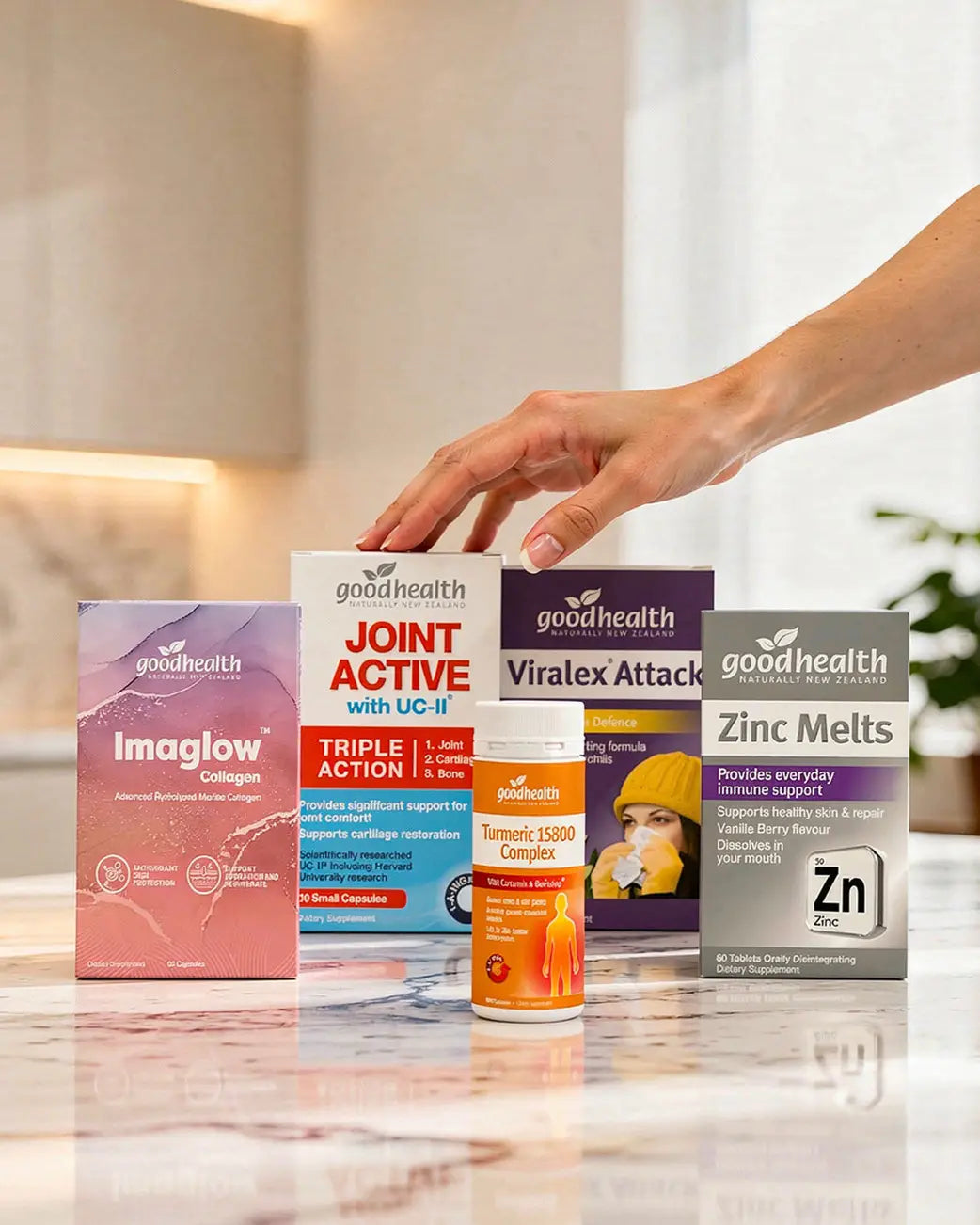 A hand reaches toward a lineup of Good Health supplement boxes and a bottle on a glossy marble countertop in a bright kitchen setting. The products include Imaglow Collagen, Joint Active with UC-II, Viralex Attack, and Zinc Melts.