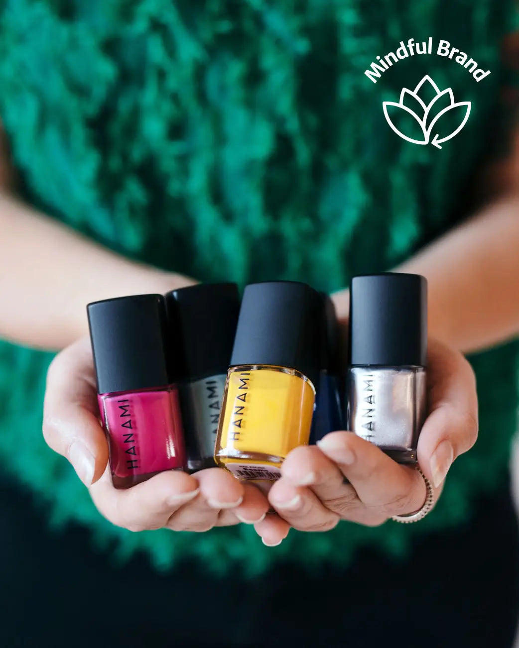 Four nail polish bottles from the brand HANAMI are held in two hands. The bottles contain pink, black, yellow, and silver nail polish, with the brand name printed vertically on the bottle.