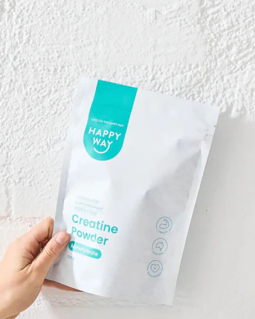 A hand holding a white stand-up pouch of Happy Way Creatine Powder against a textured white wall.