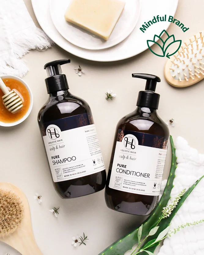 Two bottles of Holistic Hair Pure Shampoo & Conditioner on a background with honey, soap, aloe vera and body brush.