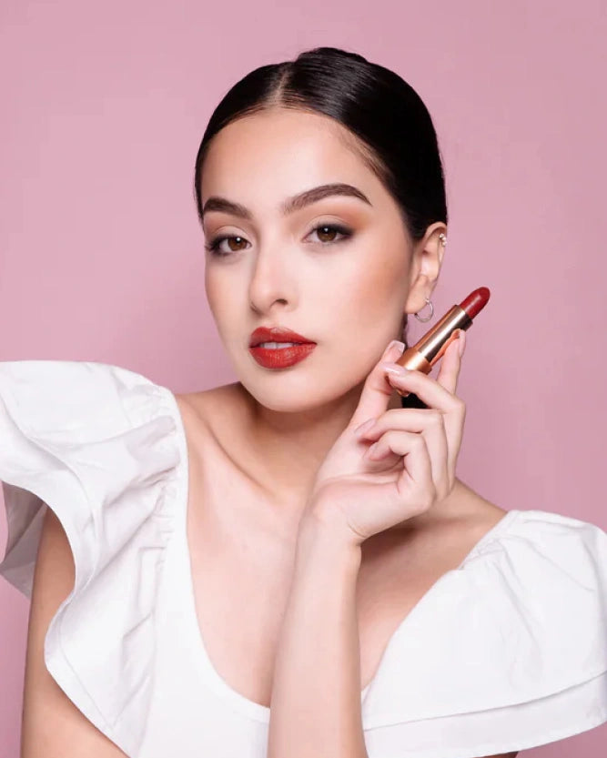 Model standing against a pink background with red lipstick on and Karen Murrell lipstick in hand.