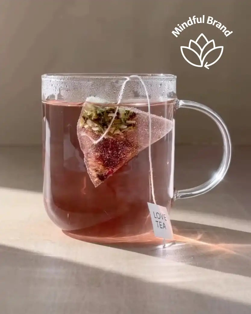Clear mug with pink tea and a triangle-shaped tea bag labeled 'Love Tea' on a neutral background, featuring the Mindful Brand logo.