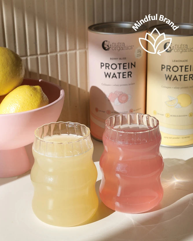 Two ribbed glass tumblers filled with pale yellow and pink protein water, placed in front of two canisters of Nutra Naturals protein water on a light surface. A pink bowl with lemons sits to the left, and a 'Mindful Brand' logo appears in the upper right.
