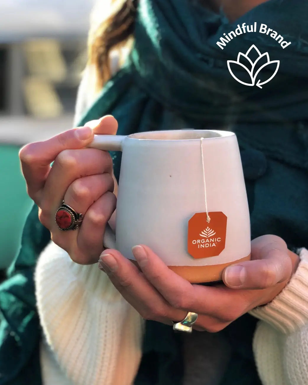 A person holding a light beige ceramic mug with a tea bag hanging from it. The mug has a terracotta-coloured tag that reads 'Organic India.'
