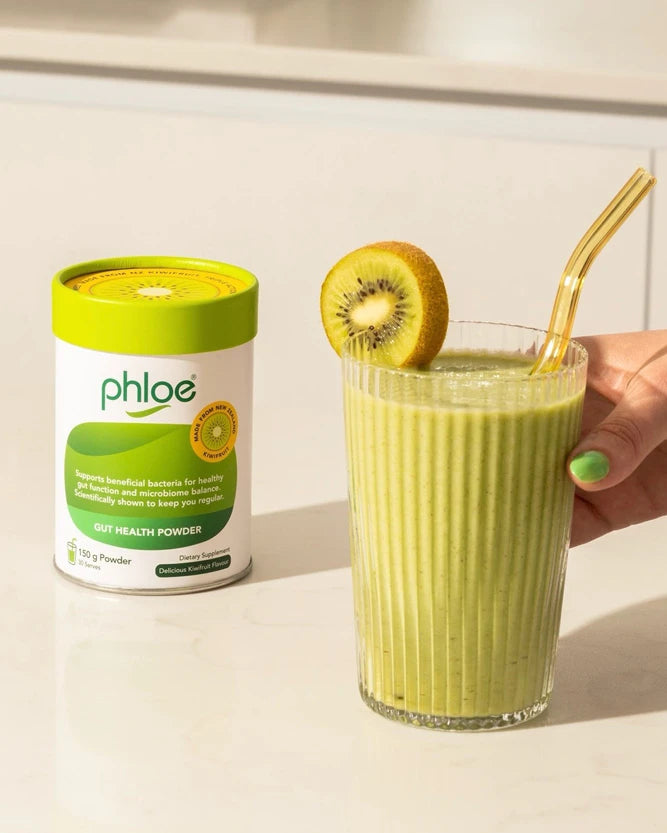 A glass of green smoothie with a gold straw and kiwi slice garnish, next to a white and green Phloe powder container on a light countertop.