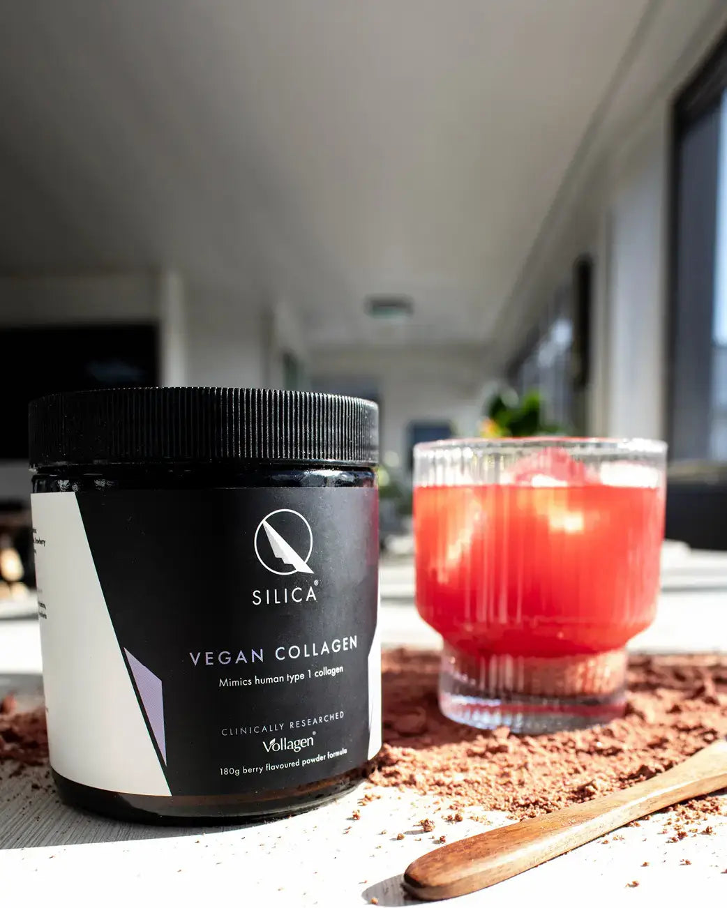 Silica Vegan Collagen powder jar in black and white packaging, shown next to a glass of pink collagen drink with a wooden spoon and scattered powder on a table in a bright indoor setting.