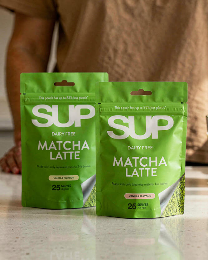 Two green 'SUP' matcha latte packages on a wooden surface with a blurred background.