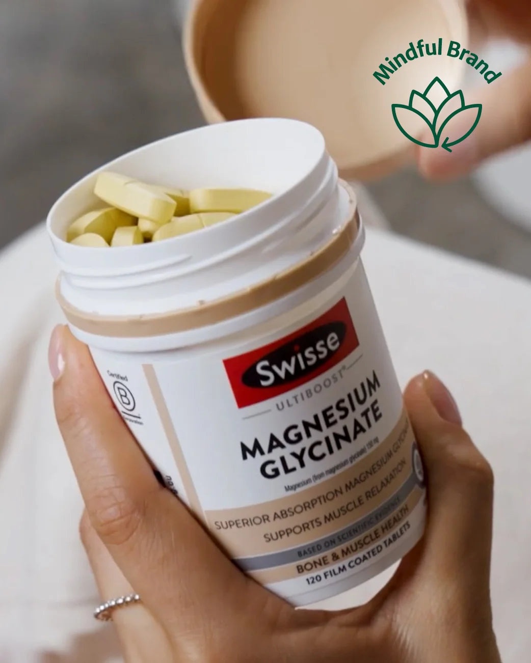 Close up of a hand holding an open bottle of Swisse Ultiboost Magnesium Glycinate with large yellow tablets visible within. Green mindful brand logo.