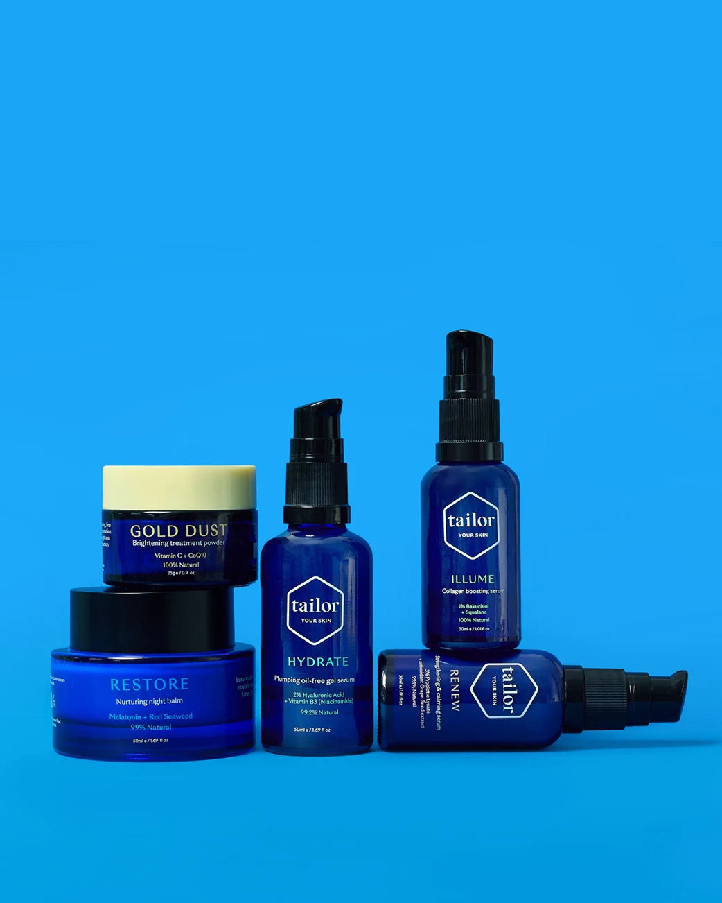 A set of blue and black personal care products from the brand tailor, including a jar of Gold Dust, a restore cream, a hydrate gel, a lumine spray, and a body spray, arranged on a solid blue background.