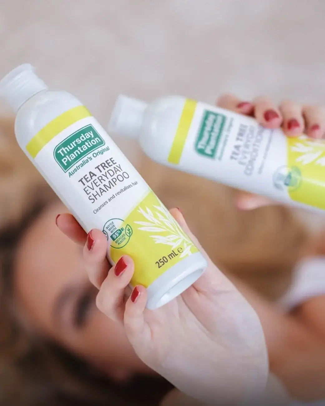 A person holds two bottles of Thursday Plantation Tea Tree Everyday Shampoo and conditioner, with one bottle in sharp focus in the foreground and the other blurred in the background.