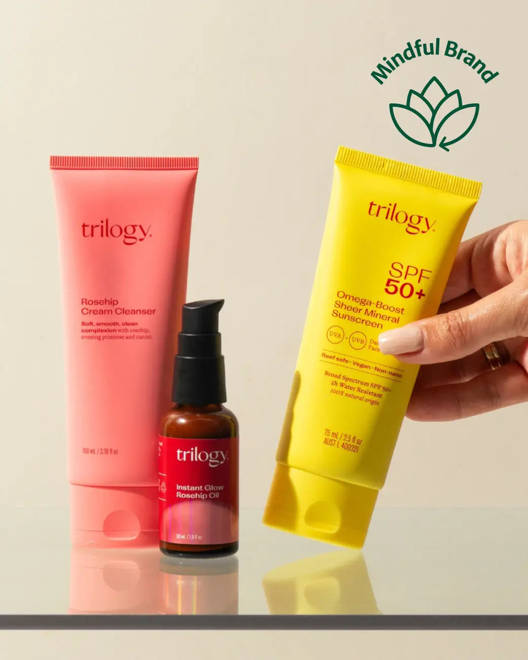 Three Trilogy skincare products are displayed on a reflective surface against a light beige background: a pink Rosehip Cream Cleanser tube, a small amber bottle with a black pump, and a yellow SPF 50+ mineral sunscreen tube. A hand holds the yellow tube, and a green 'Mindful Brand' logo appears in the top right corner.