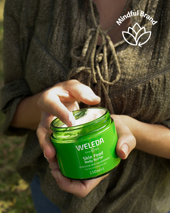 Person holding a Weleda Skin Food Body Butter jar with 'Mindful Brand' logo above.