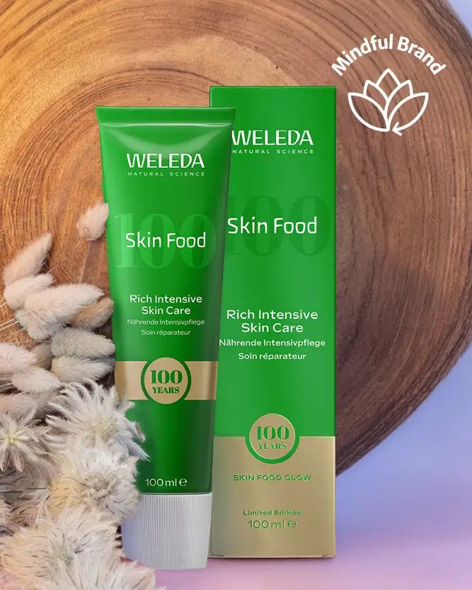 A 100ml tube and box of Weleda Skin Food Rich Intensive Skin Care, presented on a styled background with a wooden slice, dried flowers, and a purple gradient floor. A 'Mindful Brand' logo is visible in the top right corner.