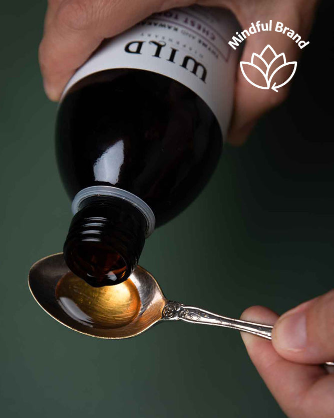 Hand pouring dark liquid from a bottle labeled 'WILD' onto a spoon with 'Mindful Brand' logo.