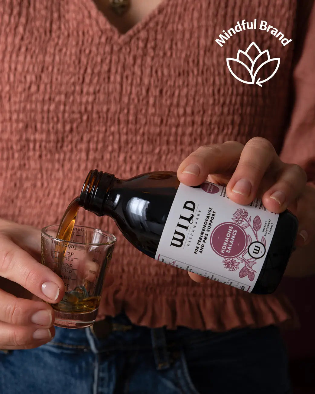 Person wearing rouched terracota top and pouring Wild Dispensary Hormone Balance into a measuring glass.